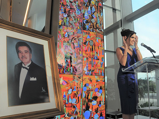 Woman clapping her hands, she's standing in front of a podium standing next to an image of a man in a suit