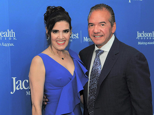 A man and woman smiling at the camera, they are standing in front of a blue step and repeat