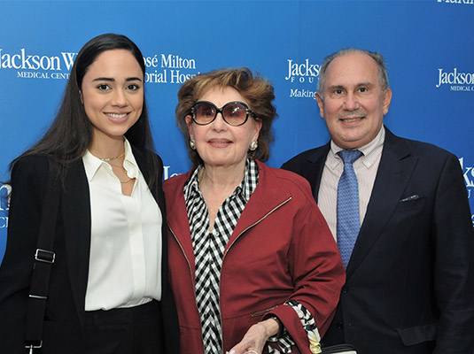 Two woman and a man standing in front of a blue step and repeat, they are all smiling at the camera