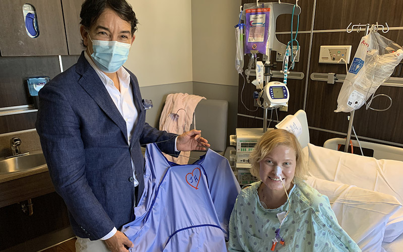 A man wearing a face mask, a suit jacket, a white dress shirt, and khaki pants is holding up a blue gown next to a woman who is smiling and sitting on a hospital bed, they both are looking at the camera