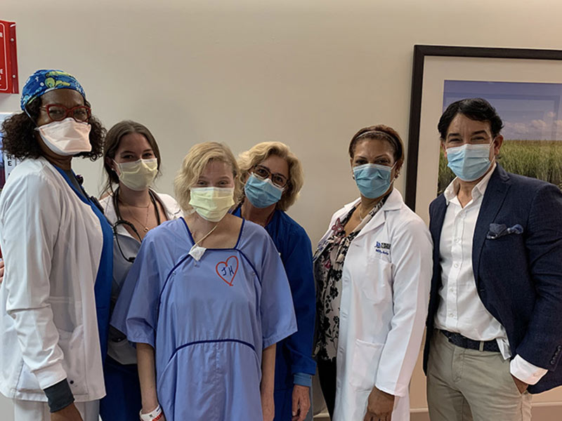 A group of medical professionals, a man, and a patient within a hospital hallway. They are all wearing face coverings and looking at the camera