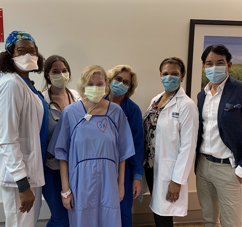 A group of medical professionals, a man, and a patient within a hospital hallway. They are all wearing face coverings and looking at the camera