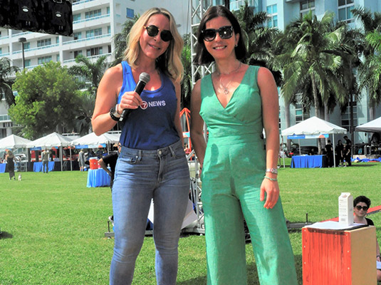 Two woman looking at the camera, one is holding a microphone, they are standing outdoors