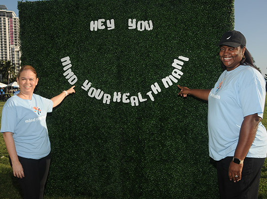 Two women pointing at a sign that reads hey you mind your health Miami