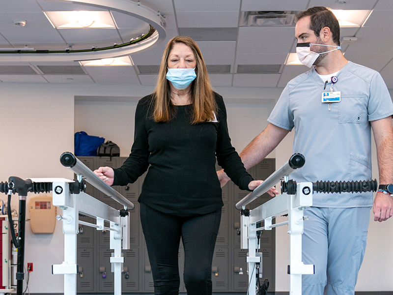 A closeup of a woman wearing black clothing and a face mask, she is walking with the help of a medical professional, she has her hands near two bars