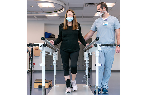 A closeup of a woman wearing black clothing and a face mask, she is walking with the help of a medical professional, she has her hands near two bars