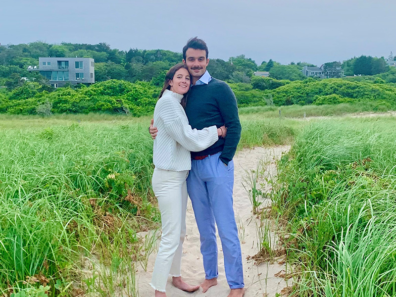 A man and woman hugging on a beach, they are looking into the camera, they are wearing sweaters and pants