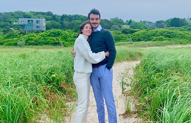 A man and woman hugging on a beach, they are looking into the camera, they are wearing sweaters and pants