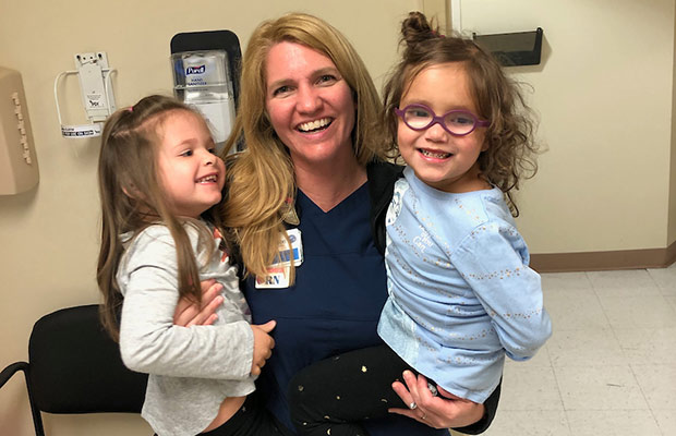 A nurse smiling at the camera, she is wearing blue scrubs and holding two young girls who are smiling