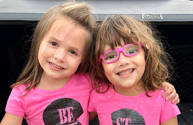 Two young girls wearing pink shirts, they smile at the camera, one young girl is wearing pink glasses