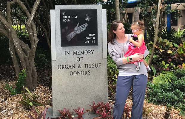 A woman holding a young girl, they stand next to a memorial that reads In memory of organ and tissue donors, in their last hour they gave a lifetime