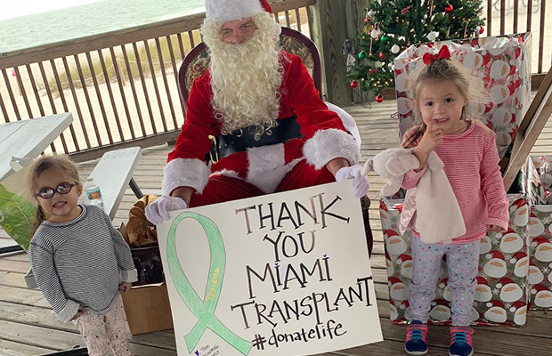 Two young girls standing next to Santa Claus, there is a sign that reads Thank You Miami Transplant, hashtag donatelife