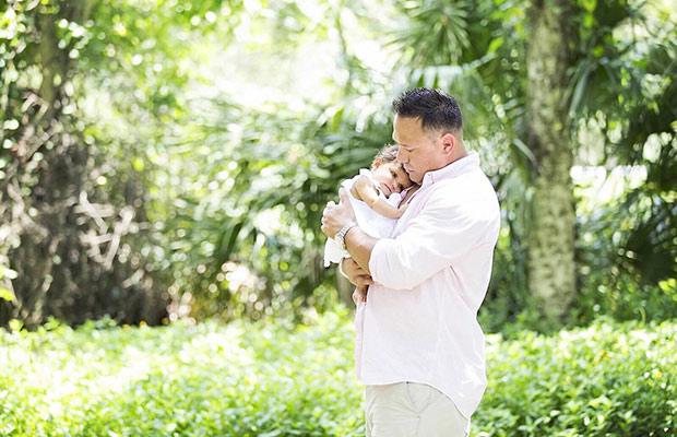 A dad holding his young daughter in his hands, they are outdoors surrounded by a lush green landscape