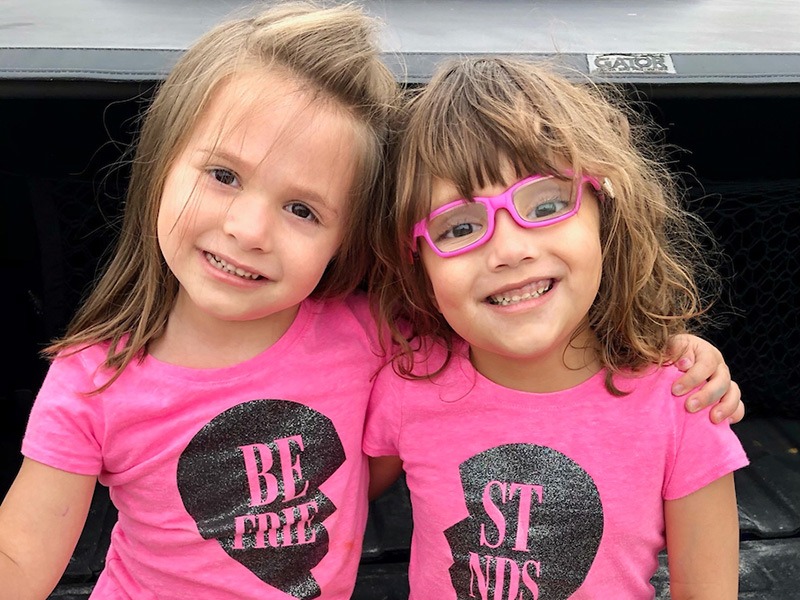 Two young girls wearing pink shirts, they smile at the camera, one young girl is wearing pink glasses