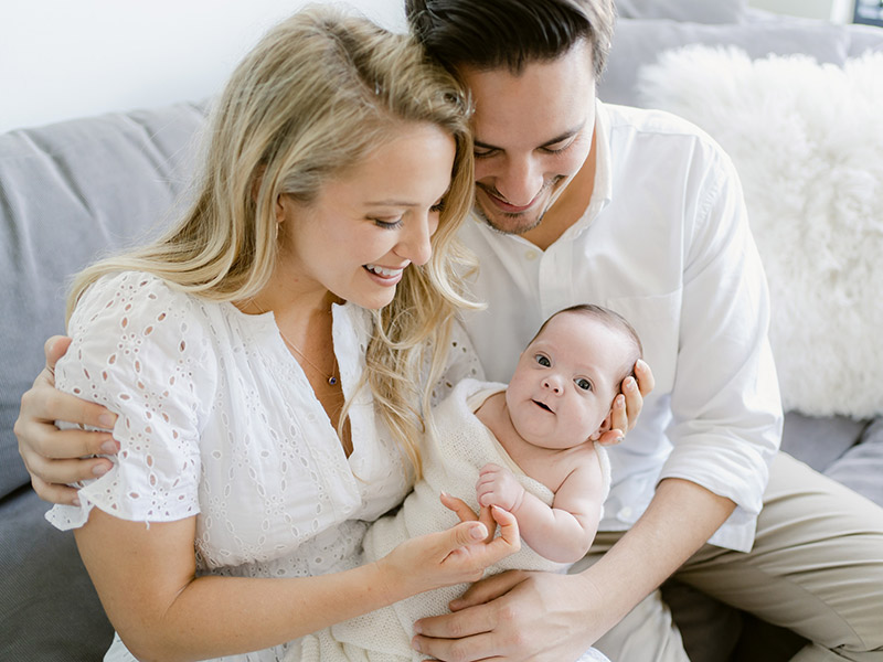 New parents holding their baby, the baby is looking at the camera, the parents are looking down and smiling at their baby