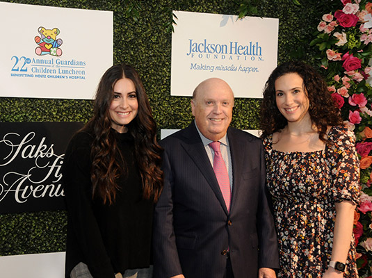 Two women and a man standing in front of a step and repeat, they smile at the camera