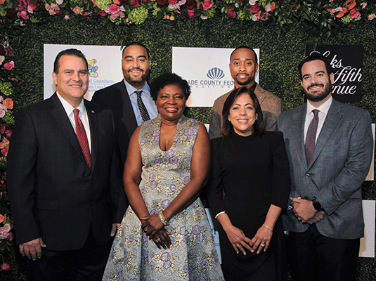 A group of six people standing in front of a step and repeat, they all smile at the camera