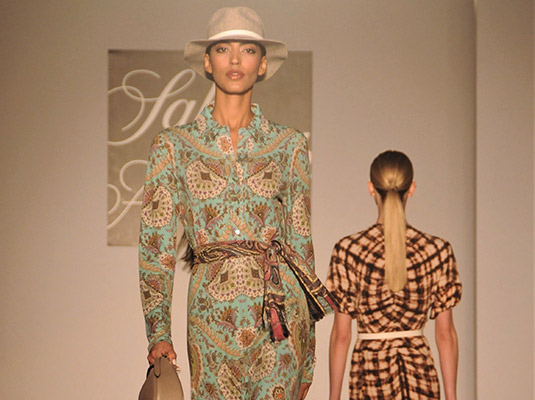 A model walking down a runway, she has on a long dress, a hat, and is holding a purse