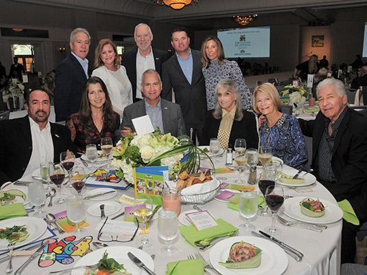 A group of eleven people sitting and standing around a circular table inside a large event room, they all smile at the camera