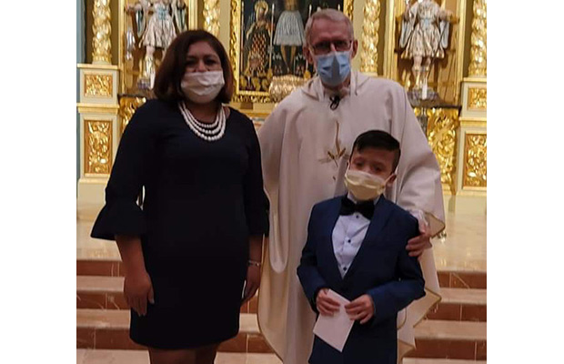 A young boy standing next to a woman and a priest, they are all looking towards the camera, they are standing inside a church