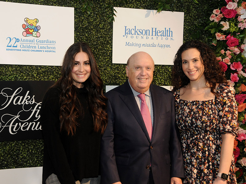 Two women and a man standing in front of a step and repeat, they smile at the camera