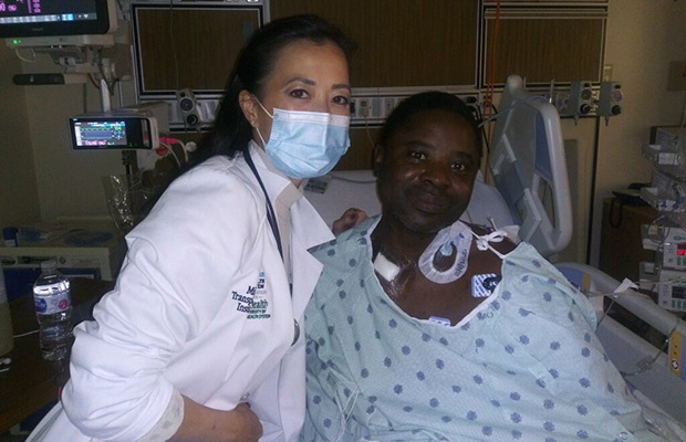 A man wearing a hospital gown, he is on a hospital bed and next to him is a medical professional, they both are looking at the camera