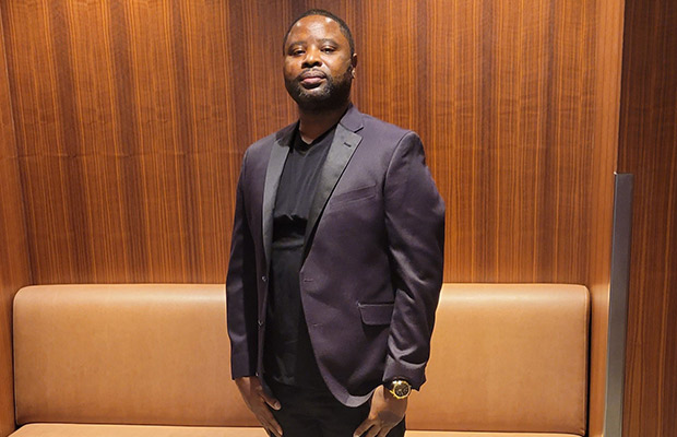 A closeup of a man, he is wearing a jacket, dark black shirt, and black slacks. He is looking at the camera.