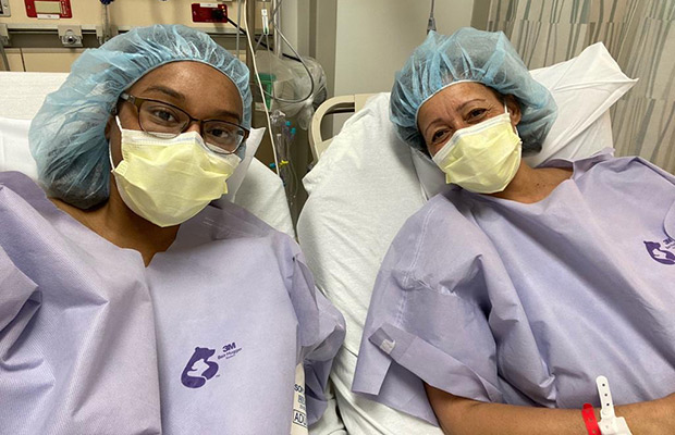 Two women laying on separate medical beds, they look at the camera, they have on scrubs and masks