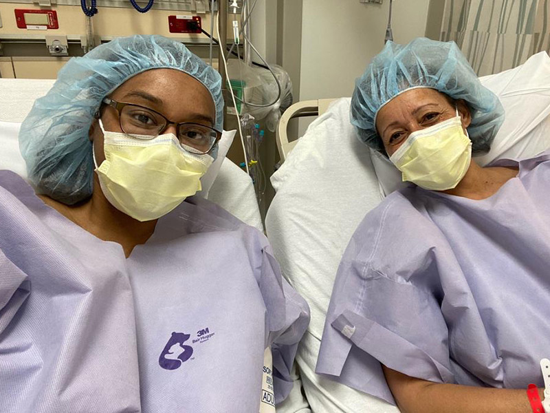 Two women laying on separate medical beds, they look at the camera, they have on scrubs and masks