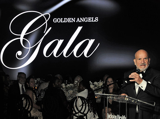A man speaking at a podium, there are people sitting behind him, there's a sign behind him that reads Golden Angels Gala