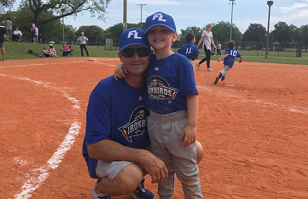 A man and his young child, they are on a baseball field and dressed in baseball attire