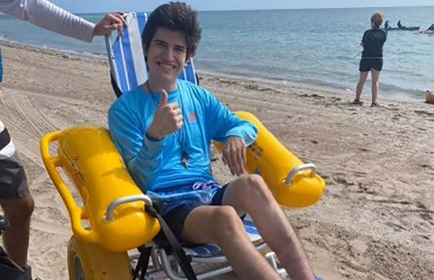 A young man laying on a beach wheelchair, he looks at the camera and has his right thumb up