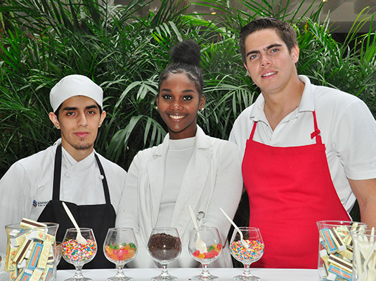 Three people standing next to each other, they stand in front of a variety of ice cream toppings