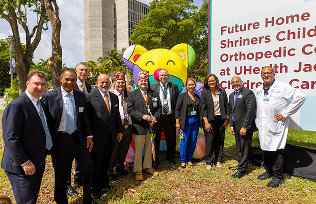 A group of eleven people standing outdoors, they are standing in front of a large colorful bear, a sign, and building