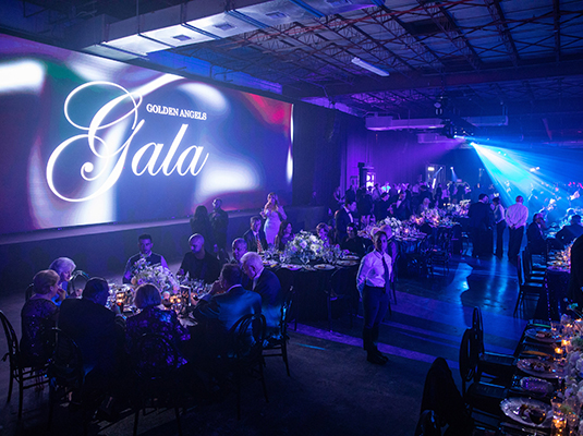 An event room filled with people, therae's a large screen that reads Golden Angels Gala