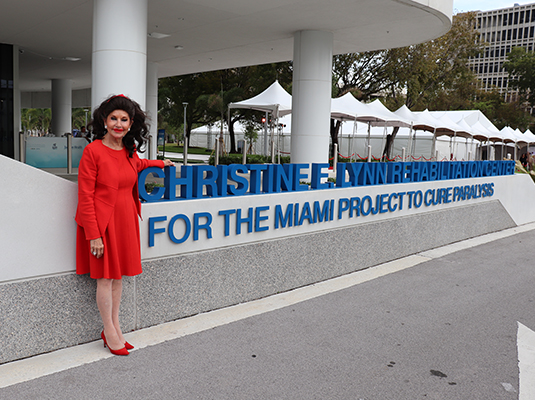 A woman standing in front of a sign that reads Christine E. Lynn Rehabilitation Center for the Miami Project to Cure Paralysis