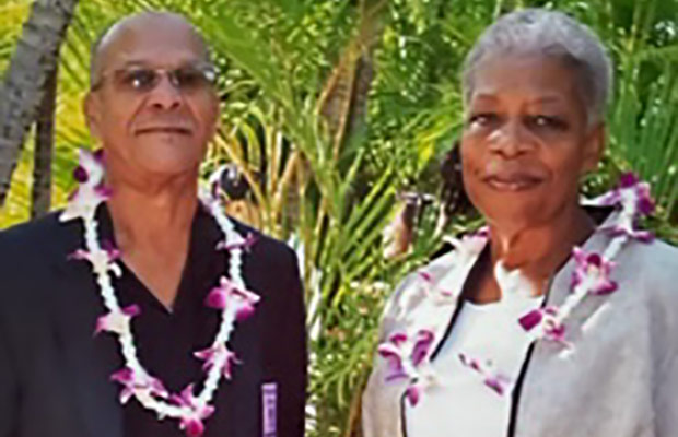 A man and women wearing flower leis, they stand outdoors and look at the camera