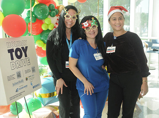 Three women standing next to a sign that reads Holiday Toy Drive