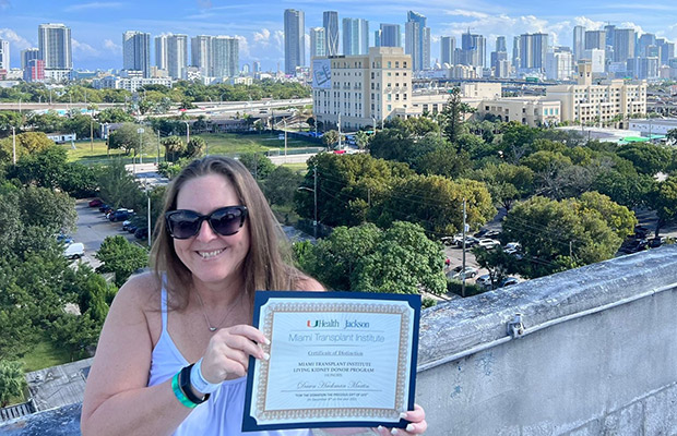 A woman holding a certificate, she is smiling at the camera and is standing outdoors, there's a view of Miami behind her