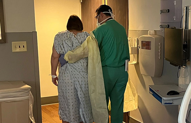 The back of a woman and medical professional, they both have gowns on, the medical professional is helping her walk