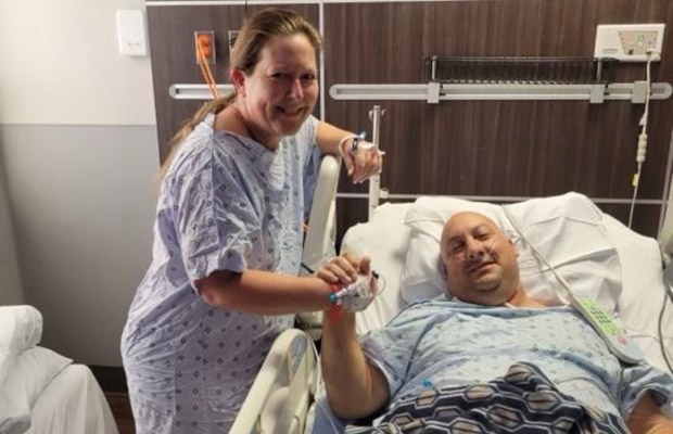 A woman and man in hospital gowns, the man is in a medical bed and the woman is standing, they hold hands and are looking at the camera