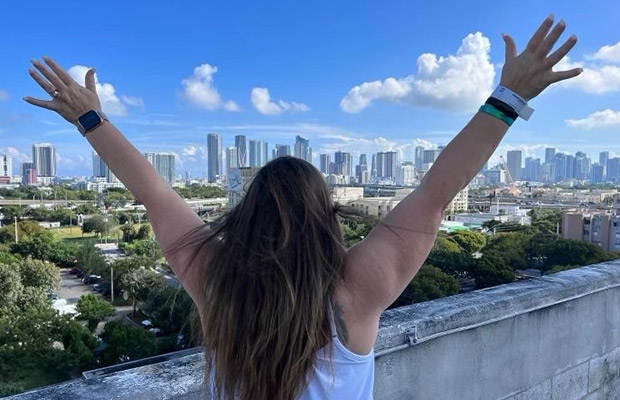 The back of a woman, she is standing outside with her arms up, there is a view of Miami in front of her