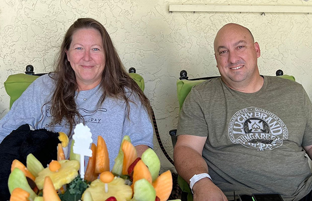 A man and woman smiling at the camera, they are sitting next to each other and there is a fruit bouquet in front of them