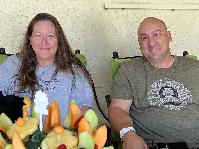 A man and woman smiling at the camera, they are sitting next to each other and there is a fruit bouquet in front of them