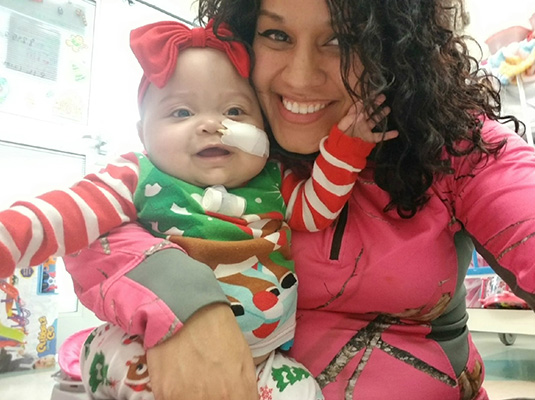 A woman and baby girl smiling at the camera, the baby has a tube coming out of left nostril
