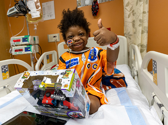 A young boy smiling at the camera, he has his left thumb up. He smiles at the camera as he opens a gift and sits on a hospital bed