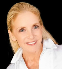 A woman with long slicked back blond hair wears a white collared shirt and smiles at the camera
