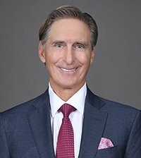 A man smiles at the camera, he has on a suit jacket, collared shirt, and maroon tie