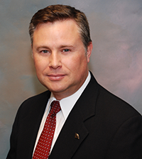 A man smiles at the camera, he has on a suit jacket, collared shirt, and red tie