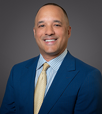A man smiles at the camera, he has on a suit jacket, collared shirt, and gold tie
